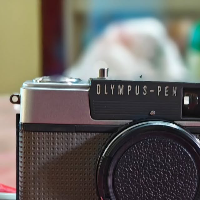 Close-up of an Olympus Pen half-frame film camera with a silver top plate and black body, lens cap on