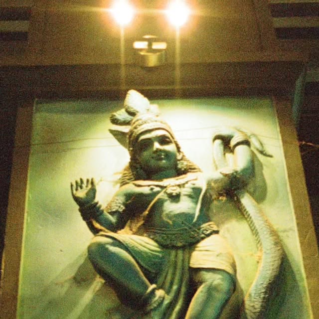 Film photograph of a temple wall sculpture of Krishna dancing on a serpent, lit by two lamps above